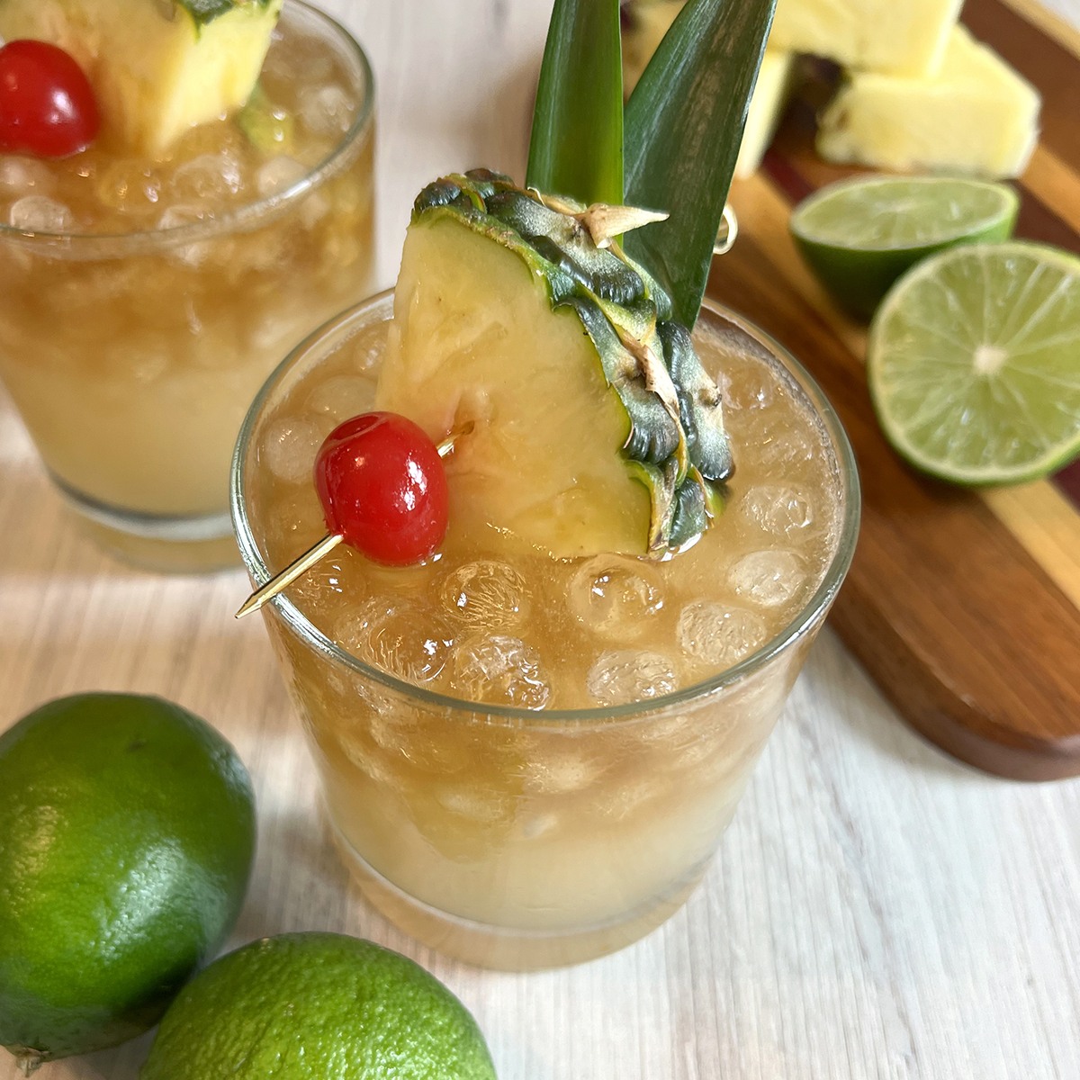 Classic mai tai served over crushed ice with a dark rum float, garnished with fresh pineapple, pineapple leaves, and a maraschino cherry, with fresh limes in the background.