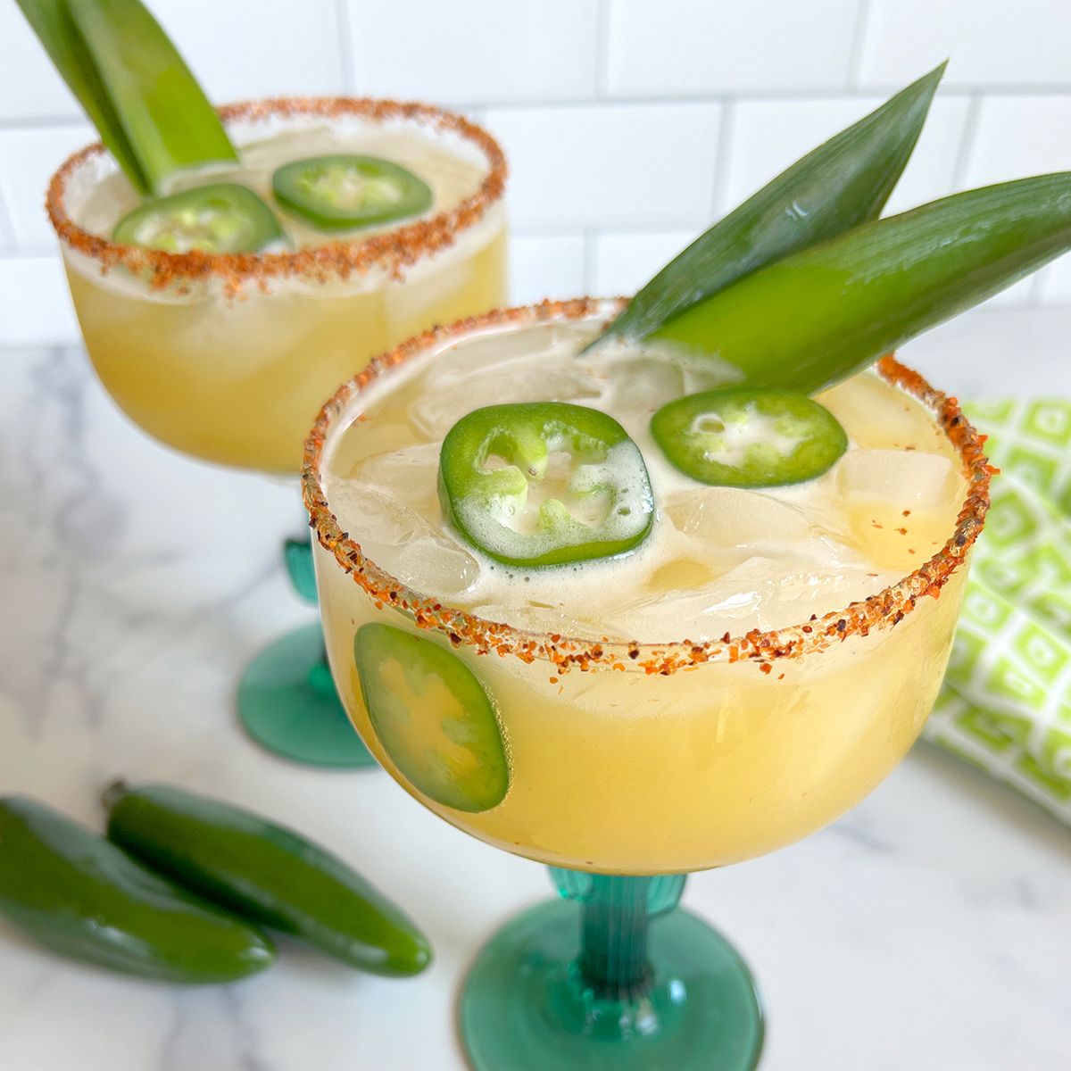 Pineapple jalapeño margaritas served over ice in glasses with Tajín-rimmed edges, fresh jalapeño slices, and pineapple leaf garnishes.